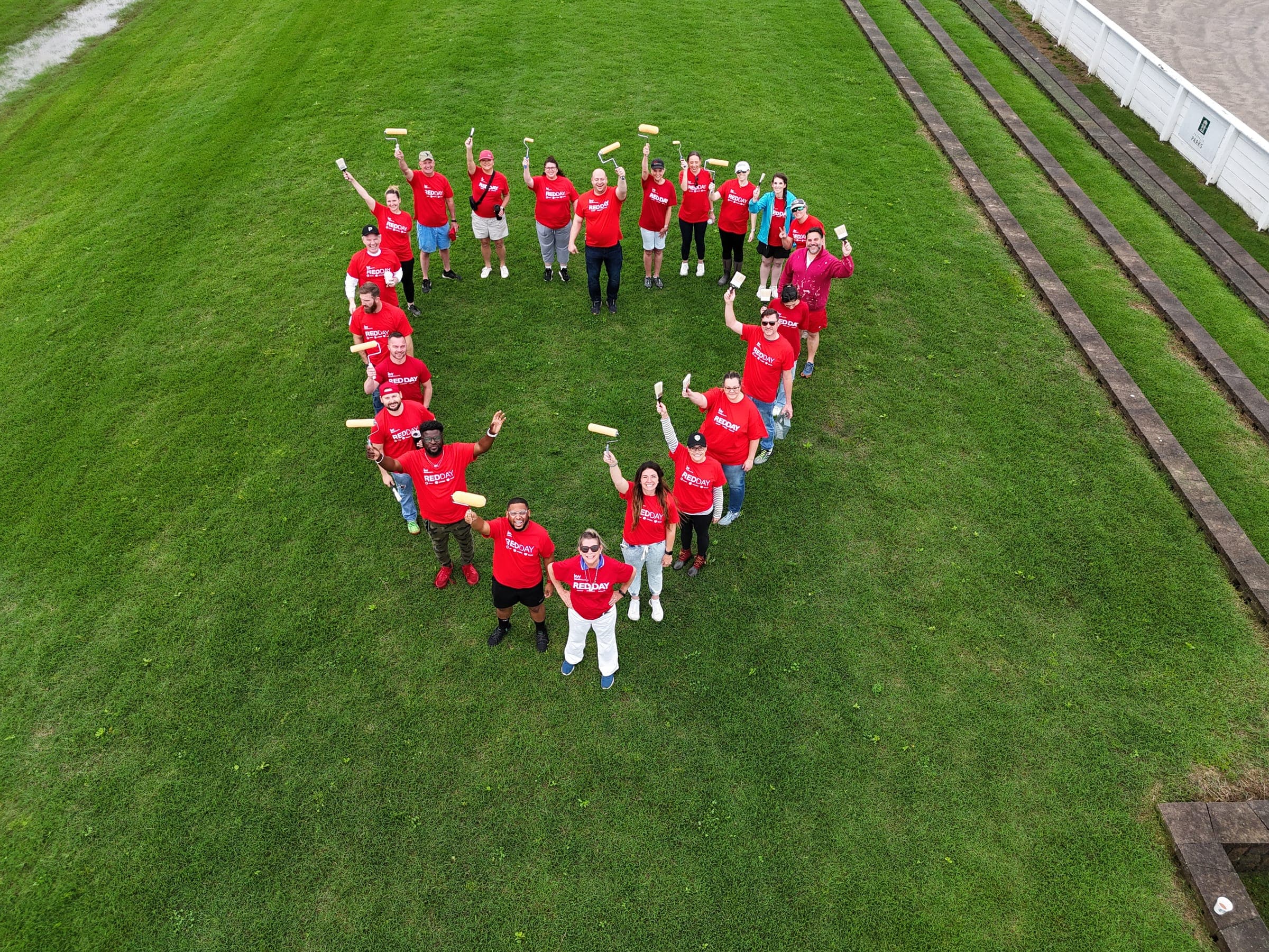 Drone view of Murfreesboro agents in red shirts forming a heart on a green field, paint rollers raised