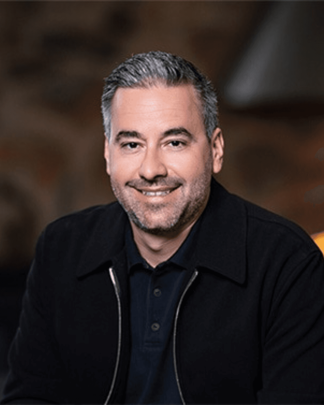 Professional headshot of Brett Tanner, a smiling man with short gray hair and salt-and-pepper stubble, wearing a black zip-up jacket over a black polo shirt, against a blurred dark brown background with a lamp visible over his shoulder.
